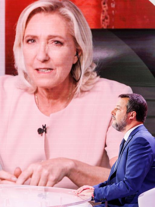 Ein Mann mit blauem Anzug sitzt in einem Fernsehstudio. Neben ihm sind Bildschirme. Darauf sind die Bilder zweier Frauen: Marine Le Pen und Ursula von der Leyen. Der Mann ist der rechte italienische Politiker Matteo Salvini.