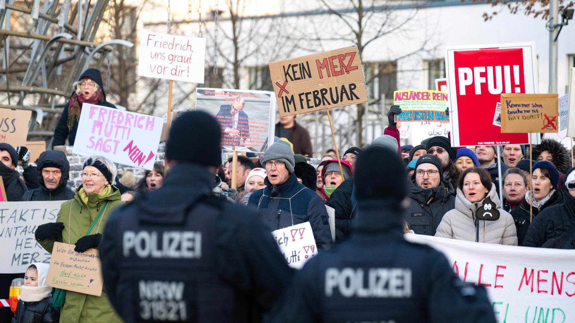 Demonstranten halten Plakate hoch und protestieren gegen die gemeinsame Abstimmung von Union und AfD. Im unscharfen Bildvordergrund stehen zwei Polizisten.