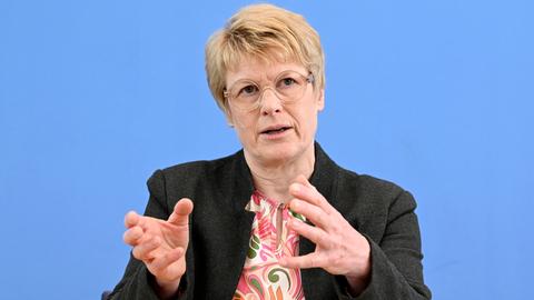 Veronika Grimm spricht im Haus der Bundespressekonferenz vor einem blauen Hintergrund. Sie trägt eine runde Brille, eine bunte Bluse und ein schwarzes Jackett.