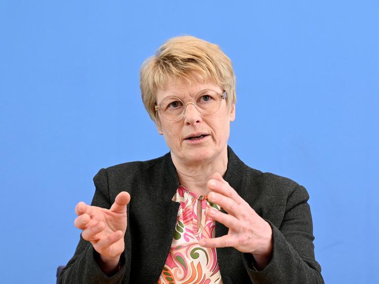 Veronika Grimm spricht im Haus der Bundespressekonferenz vor einem blauen Hintergrund. Sie trägt eine runde Brille, eine bunte Bluse und ein schwarzes Jackett.