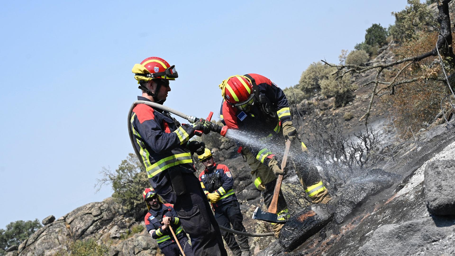 Zwei Feuerwehrleute spritzen Wasser auf einen bereits abgebrannten Bereich. Sie tragen Schutzkleidung und Helme. Die Pflanzen sind verkohlt und Felsen schwarz. Zwei Feuerwehrleute spritzen Wasser auf einen bereits abgebrannten Bereich. Sie tragen Schutzkleidung und Helme. Die Pflanzen sind verkohlt und Felsen schwarz.