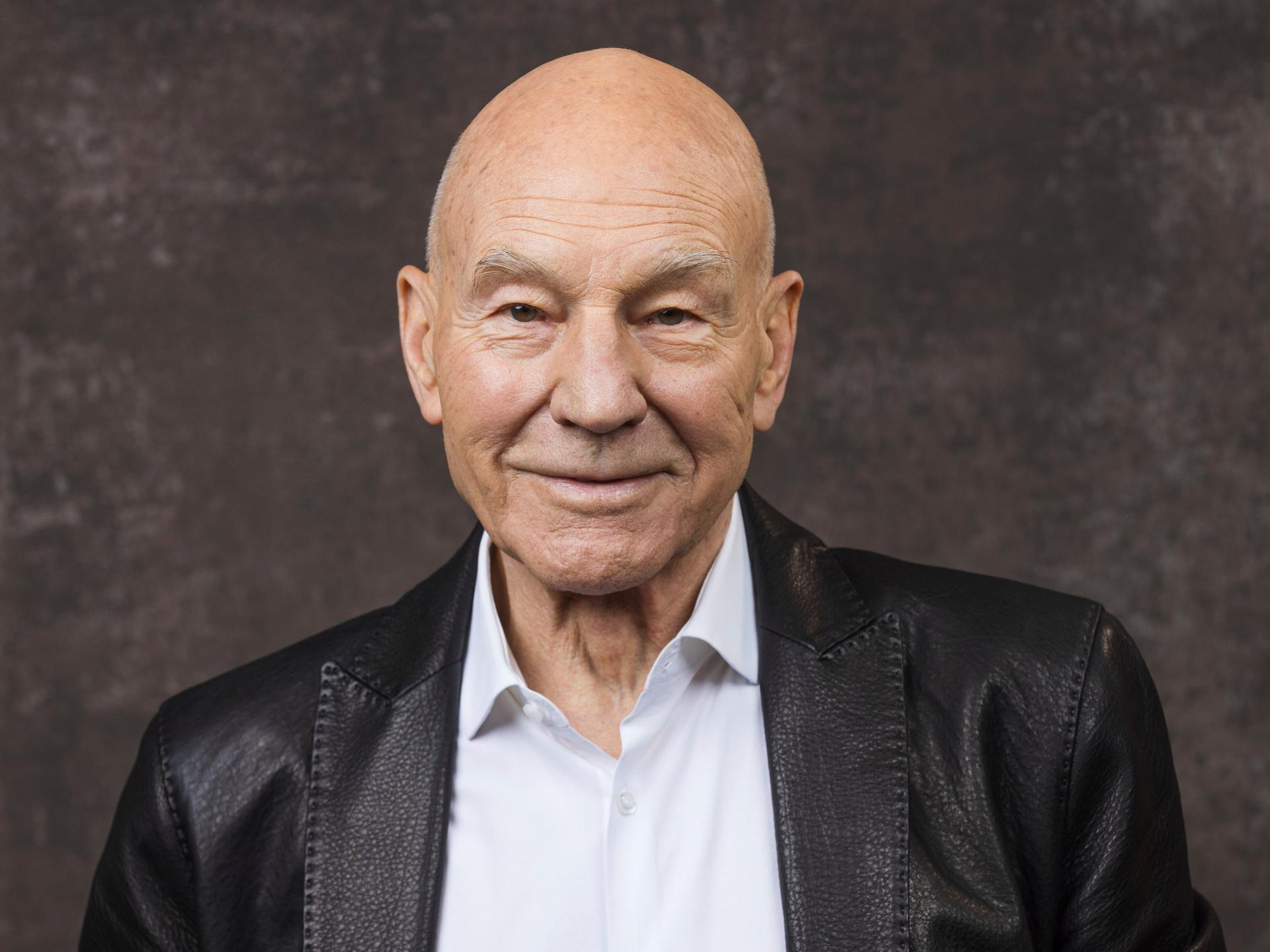 Patrick Stewart posiert in einer schwarzen Lederjacke vor neutralem Hintergrund für ein Porträt.