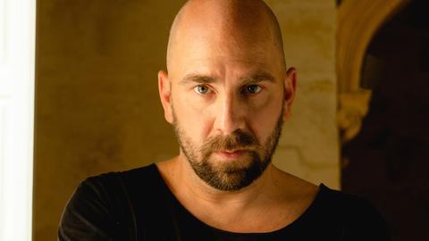 Sebastian Studnitzky, ein Mann mit Glatze und Vollbart, schaut mit ernstem Blick frontal in die Kamera. Er hat die Arme verschränkt und trägt ein schwarzes T-Shirt. Im Hintergrund ist ein altes Gemäuer und die Andeutung eines Torbogens zu sehen.