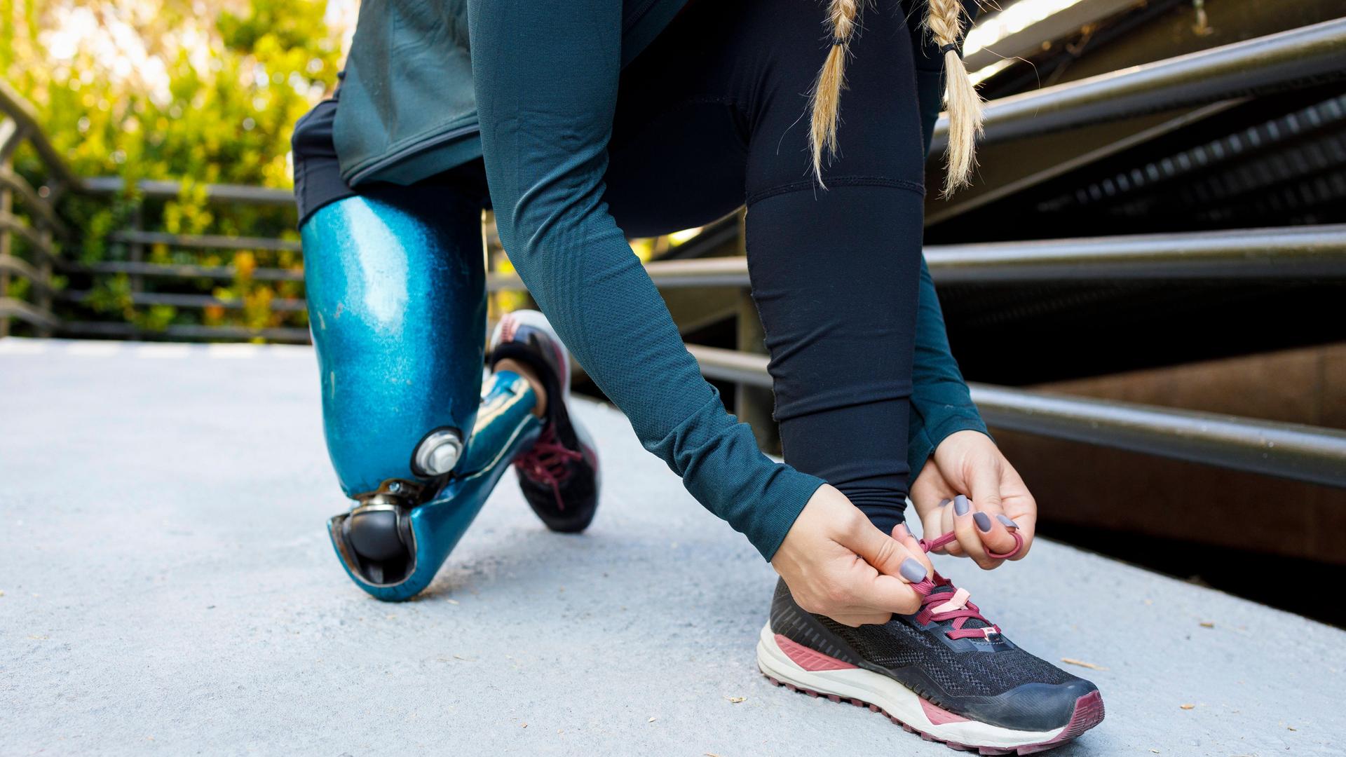 Eine Frau mit einer Beinprothese kniet auf dem Boden und schnürt ihre Joggingschuhe. (Symbolbild)