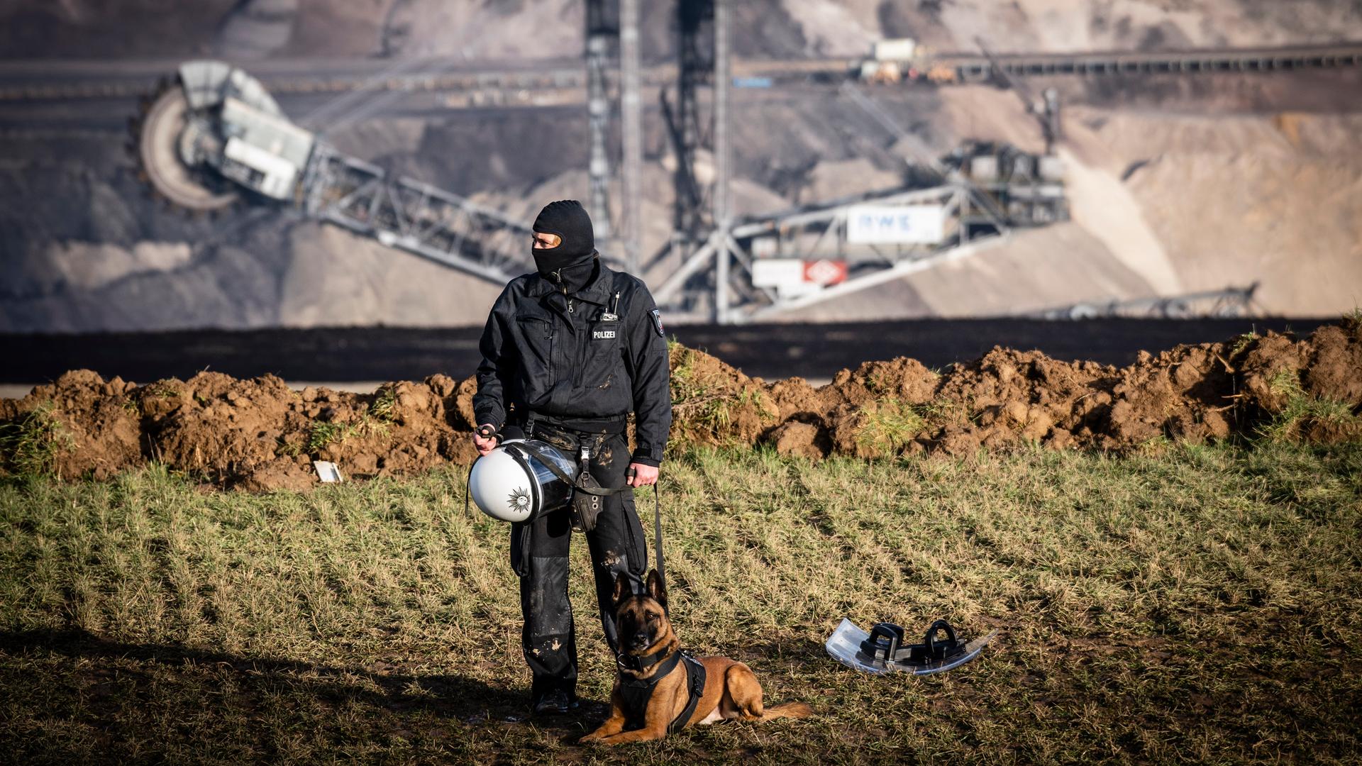 Ein Polizist in voller Montur steht zusammen mit einem angeleinten Hund am Rande des Tagebaus in Lützerath bei Erkelenz. Im Hintergrund ist ein riesiger Bagger im Tagebau zu sehen.