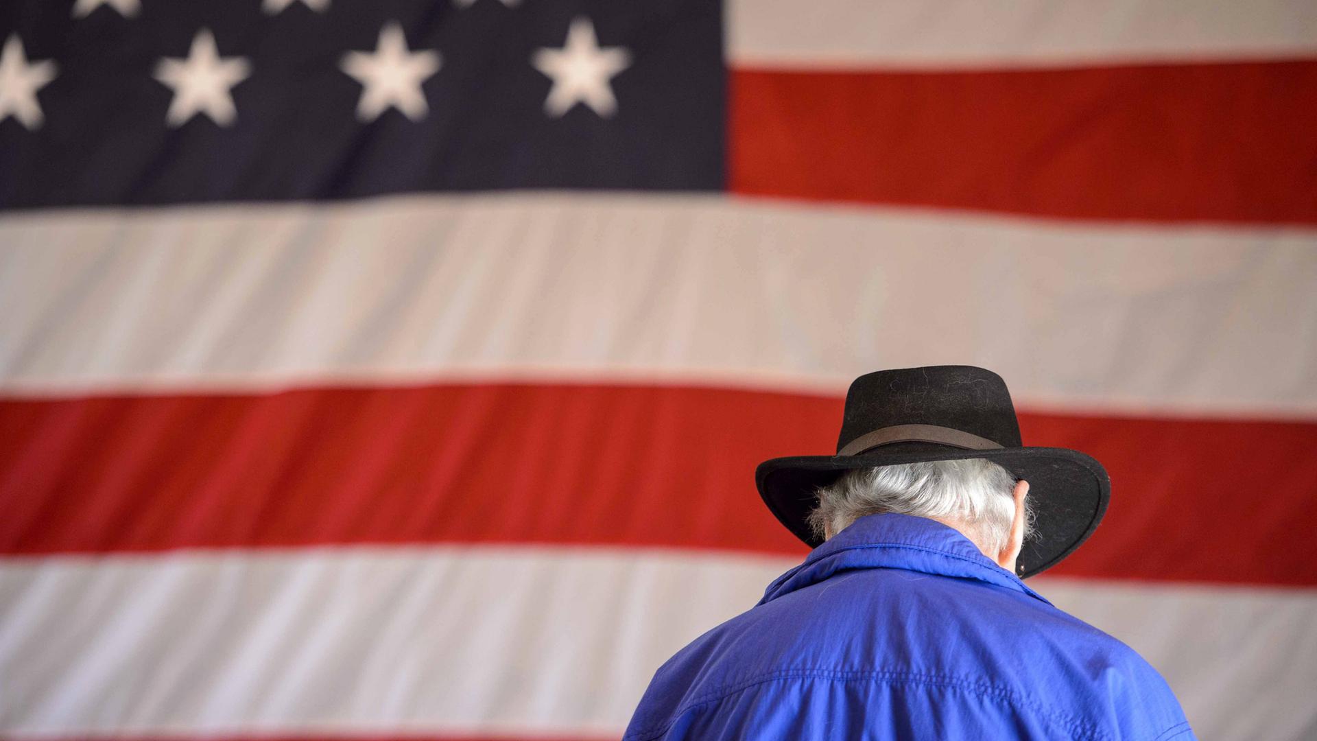 Ein Mann mit grauem Haar und Cowboyhut steht vor einer amerikanischen Flagge.