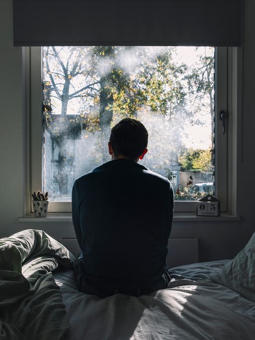 Rückenansicht eines Mannes, der auf einem Bett sitzt und aus dem Fenster schaut