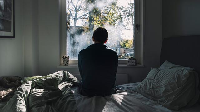 Rückenansicht eines Mannes, der auf einem Bett sitzt und aus dem Fenster schaut Rückenansicht eines Mannes, der auf einem Bett sitzt und aus dem Fenster schaut