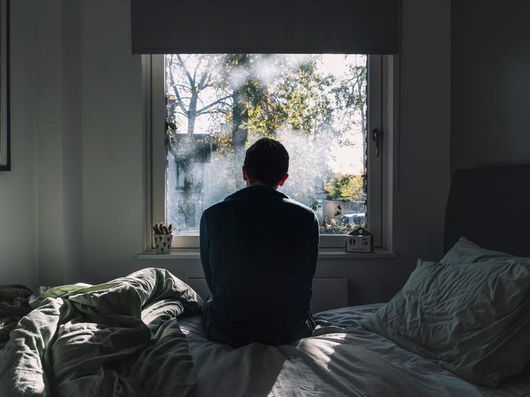 Rückenansicht eines Mannes, der auf einem Bett sitzt und aus dem Fenster schaut