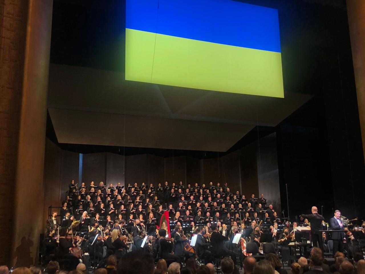 Der Blick auf die Bühne zeigt den großen, aufgestellten Chor, der unter einer ukrainischen Flagge singt.