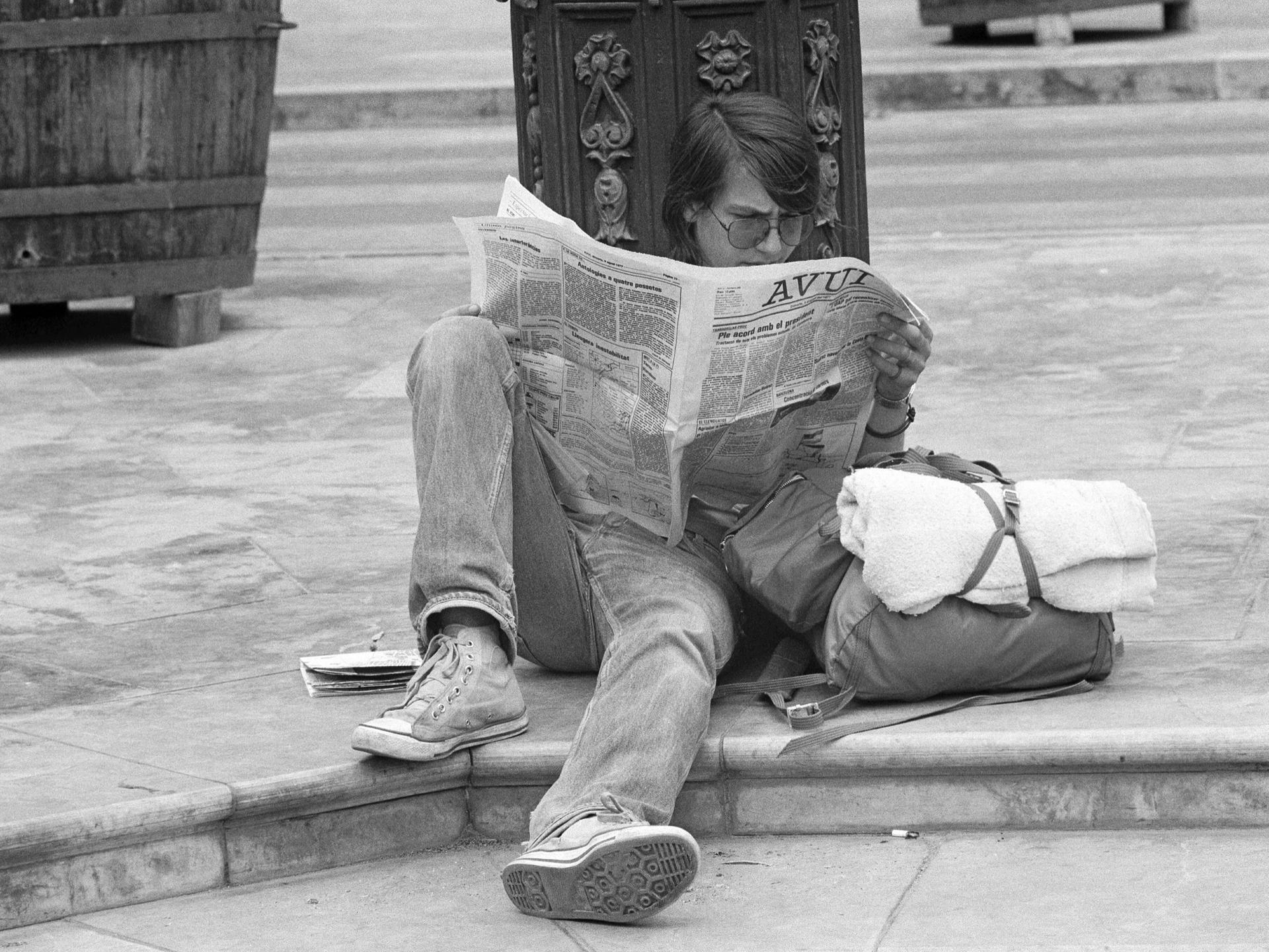 Ein Schwarz-Weiß-Foto zeigt einen Jugendlichen, der im Jahr 1977 in Valencia in Spanien am Straßenrand auf dem Boden sitzt und eine Zeitung liest.