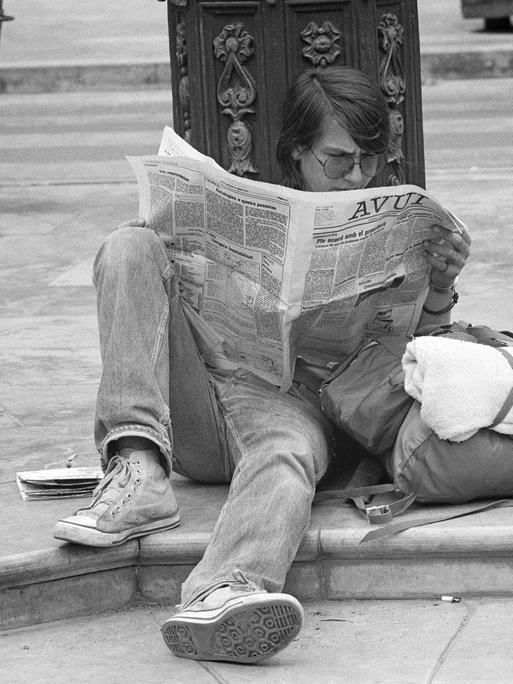 Ein Schwarz-Weiß-Foto zeigt einen Jugendlichen, der im Jahr 1977 in Valencia in Spanien am Straßenrand auf dem Boden sitzt und eine Zeitung liest.
