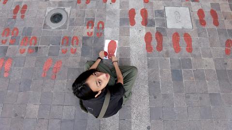 Blick auf eine Frau, die rote Schuhabdrücke auf die Strasse malt. Sie schaut nach oben in die Kamera.