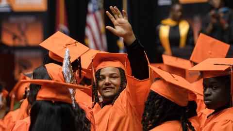 Studenten in orangener Kleidung bei der Abschlussfeier. Ein junger Mann reckt einen Arm empor.