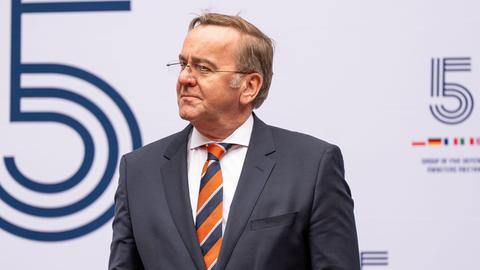 Bundesverteidigungsminister Pistorius steht  vor einer weißen Wand mit blauem Logo, er trägt eine gestreifte Krawatte.