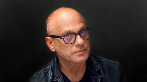 Ein Mann mit Halbglatze und schwarzer Hornbrille schaut ernst aus dem Bild hinaus. Der Hintergrund und seine Kleidung sind schwarz, seine Augen blau.
