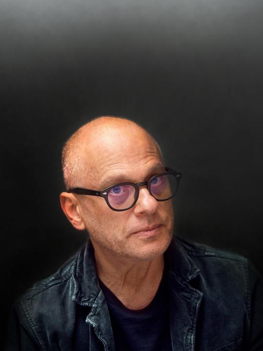 Ein Mann mit Halbglatze und schwarzer Hornbrille schaut ernst aus dem Bild hinaus. Der Hintergrund und seine Kleidung sind schwarz, seine Augen blau.