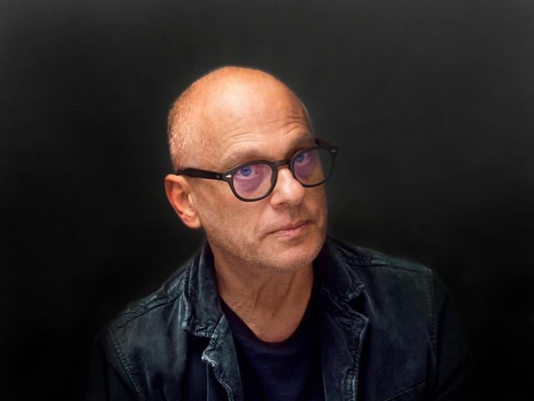 Ein Mann mit Halbglatze und schwarzer Hornbrille schaut ernst aus dem Bild hinaus. Der Hintergrund und seine Kleidung sind schwarz, seine Augen blau.