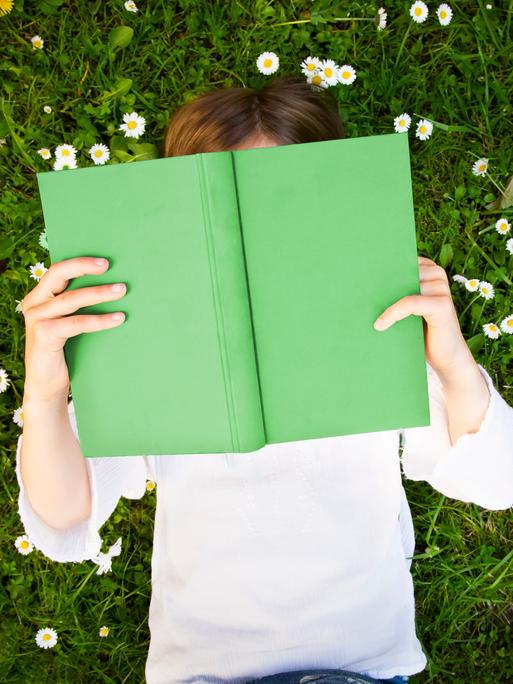 Ein junger Mensch liegt auf einer Wiese mit Gänseblümchen und hält ein grünes Buch in der Hand.