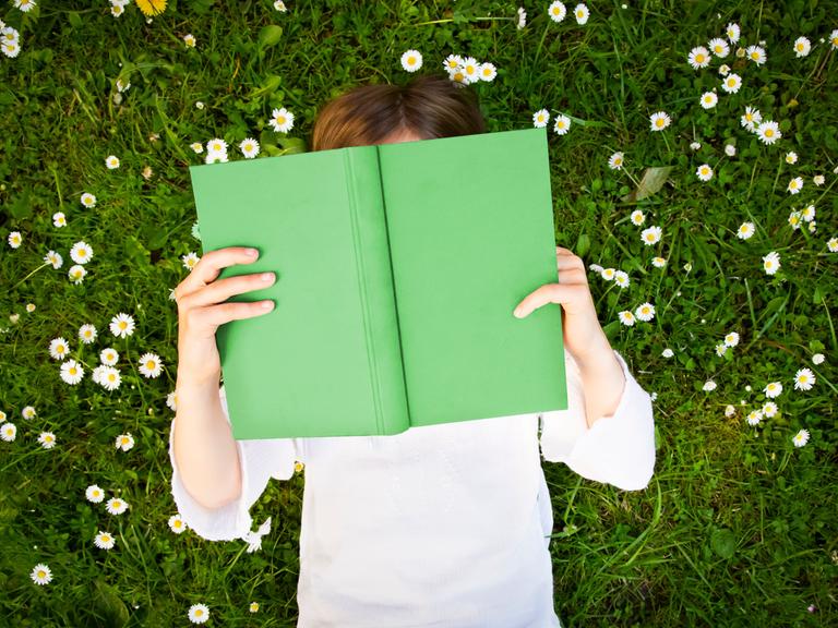 Ein junger Mensch liegt auf einer Wiese mit Gänseblümchen und hält ein grünes Buch in der Hand.