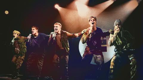 Take That stehen auf einer Bühne und führen eine Tanzchoreografie auf