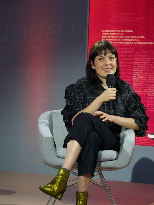 Dana von Suffrin sitzt in schwarzer Kleidung in einem grauen Stuhl auf der Buchmesse in Leipzig und spricht in ein Mikrofon.