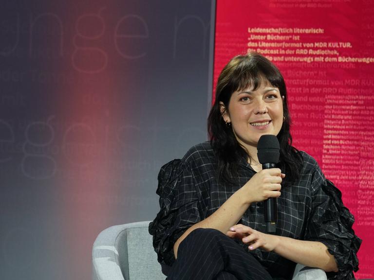 Dana von Suffrin sitzt in schwarzer Kleidung in einem grauen Stuhl auf der Buchmesse in Leipzig und spricht in ein Mikrofon.