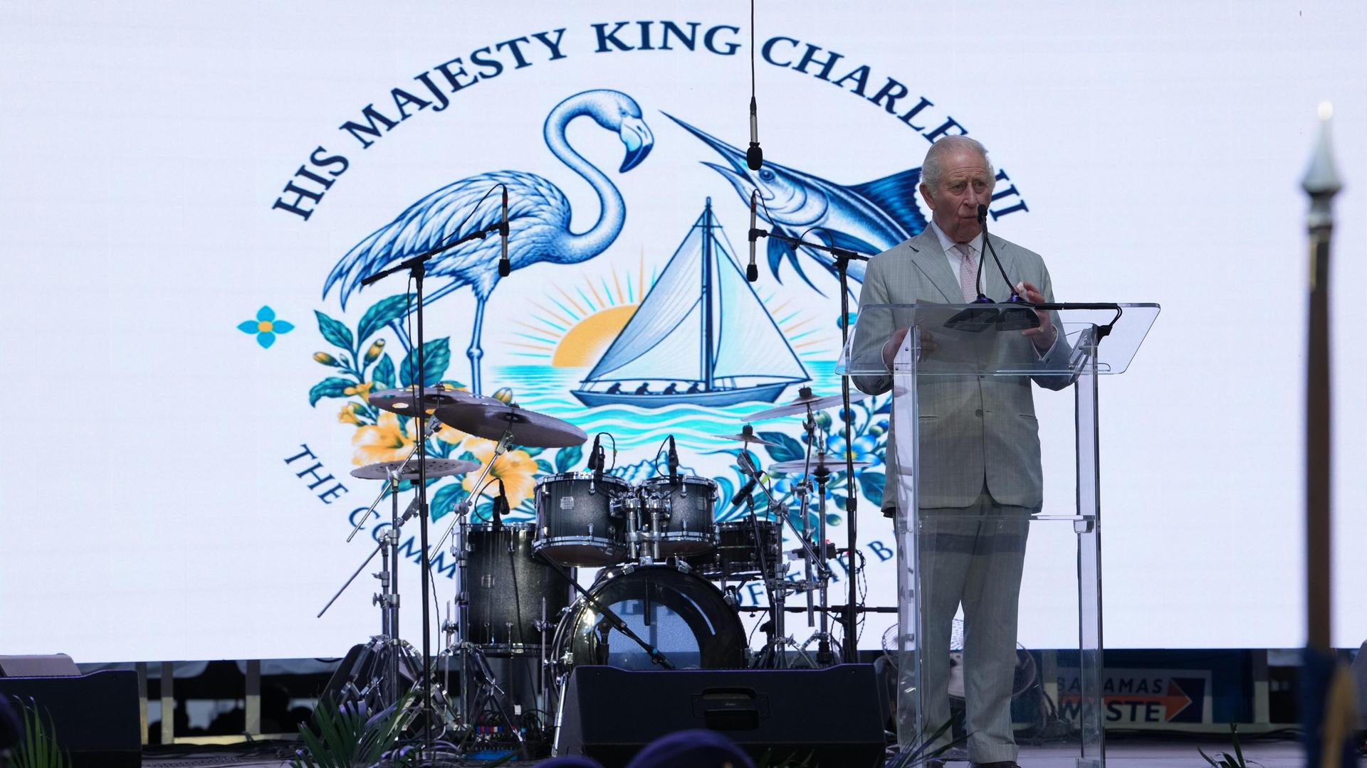 König Charles steht an einem Rednerpult, hinter ihm steht ein Schlagzeug, dahinter ein Bild mit einem Flamingo, einem Schwertfisch und einem Segelboot, das ihn ehrt.