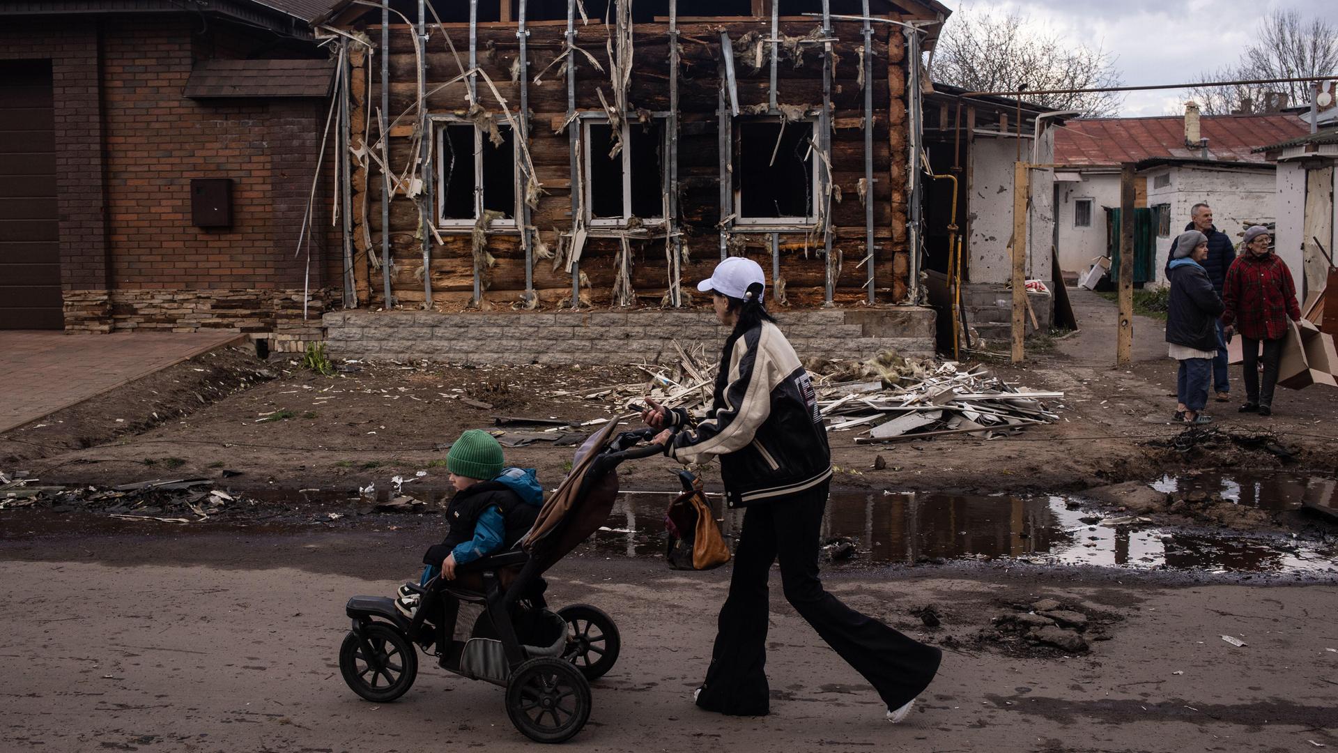Ukraine, Sumy Region: Eine Frau mit Kinderwagen geht an einem zerstörten Haus vorbei, drei Personen stehen im Hintergrund und unterhalten sich. 