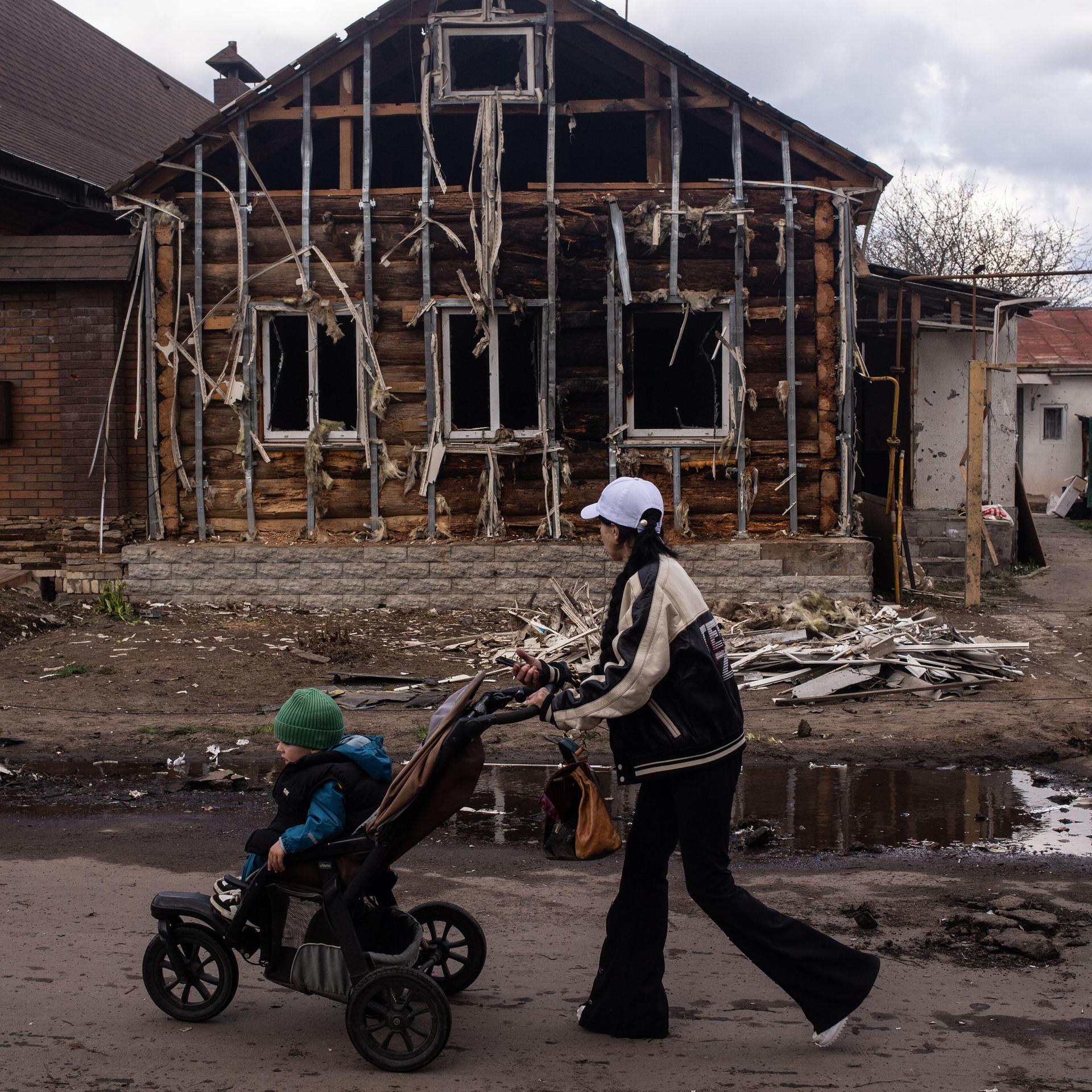 Ukraine, Sumy Region: Eine Frau mit Kinderwagen geht an einem zerstörten Haus vorbei, drei Personen stehen im Hintergrund und unterhalten sich. 