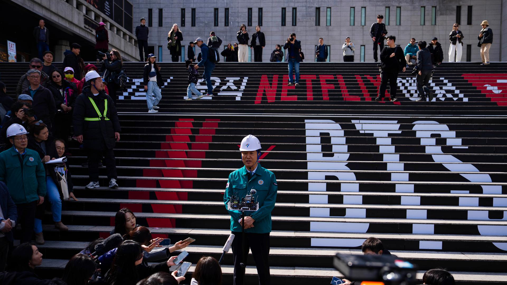 Seouls Bürgermeister gibt eine Pressekonferenz am Konzert-Ort für das BTS-Comeback auf einer Treppe. Darauf die Schriftzüge BTS und Netflix 