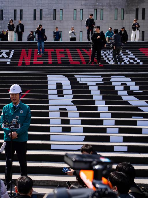 Seouls Bürgermeister gibt eine Pressekonferenz am Konzert-Ort für das BTS-Comeback auf einer Treppe. Darauf die Schriftzüge BTS und Netflix 