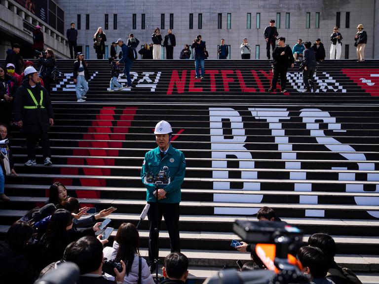 Seouls Bürgermeister gibt eine Pressekonferenz am Konzert-Ort für das BTS-Comeback auf einer Treppe. Darauf die Schriftzüge BTS und Netflix 