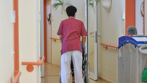 Ein Pfleger ist von hinten zu sehen, wie er einen Pflegebedürftigen im Rollstuhl schiebt.