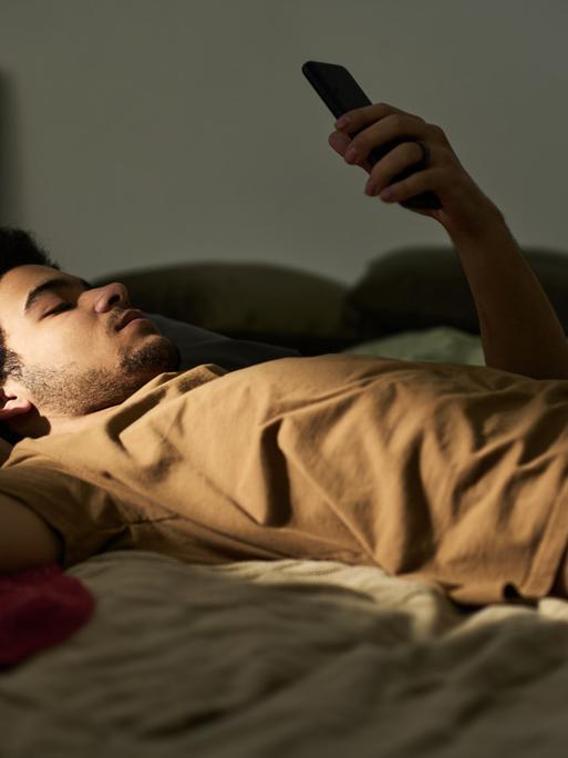 Ein Mann liegt auf dem Rücken im Bett. Sein linker Arm ist aufgestützt und er hält ein Smartphone in die Höhe, auf das er blickt.