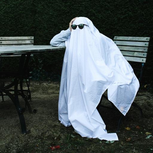Ein Gespenst mit Sonnenbrille sitzt geknickt an einem Tisch und stützt seinen Kopf ab.