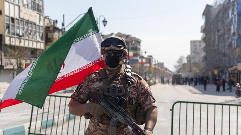 Ein Soldat der Iranischen Revolutionsgarde steht vor einer abgesperrten Straße, neben ihm weht die iranische Flagge