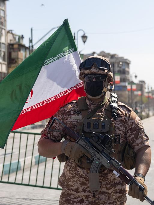 Ein Soldat der Iranischen Revolutionsgarde steht vor einer abgesperrten Straße, neben ihm weht die iranische Flagge