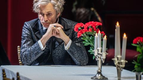 Ein Mann mit grauen Haaren sitzt an einem Tisch mit Kerzen und roten Blumen, stützt sich mit den Ellenbogen auf den Tisch und schaut nachdenklich zur Seite