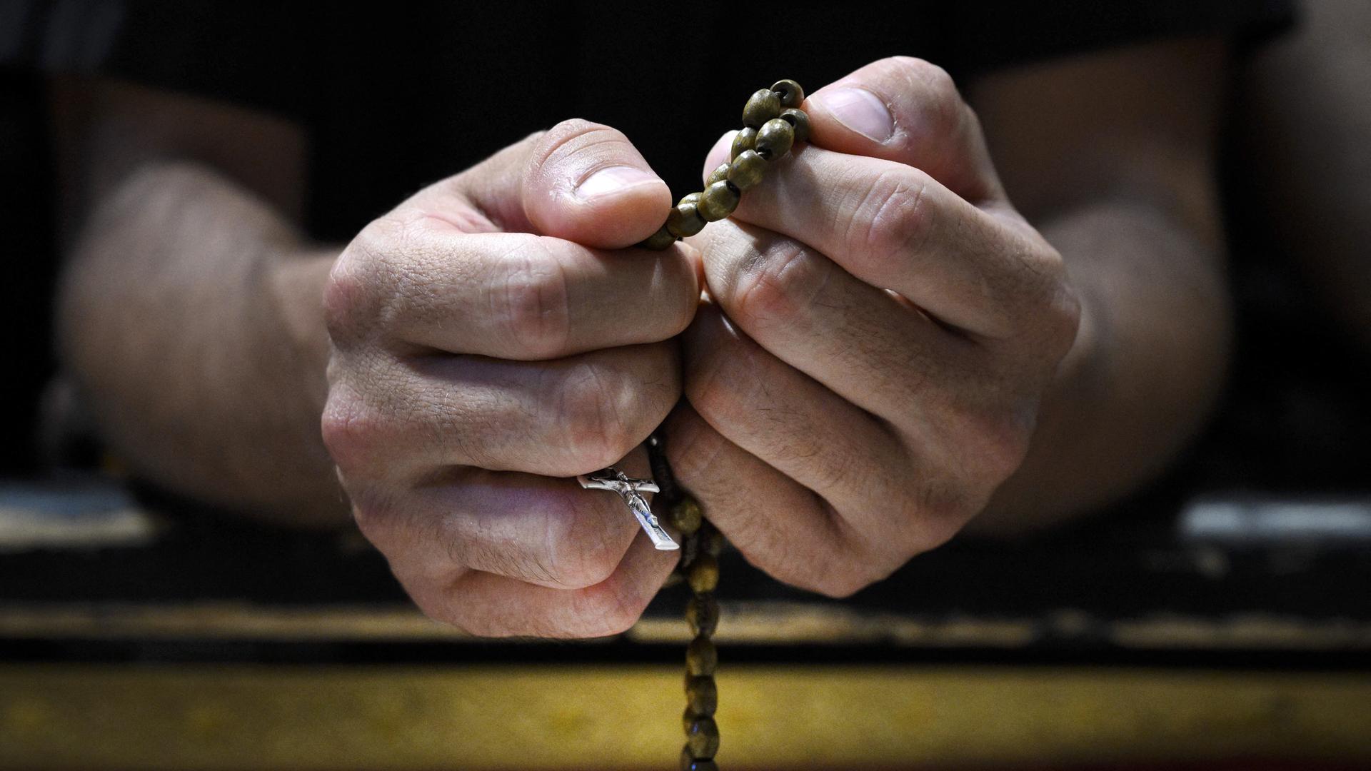 Großaufnahme zweier Hände: Ein Gläubiger hält einen Rosenkranz sehr fest. Das Kruzifix ist durch seine Finger zu erkennen.