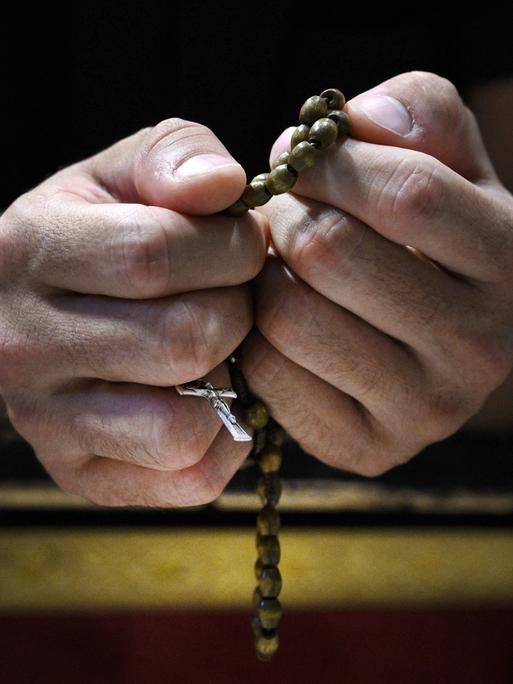 Großaufnahme zweier Hände: Ein Gläubiger hält einen Rosenkranz sehr fest. Das Kruzifix ist durch seine Finger zu erkennen.