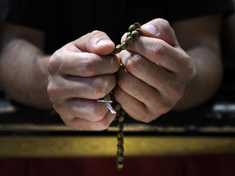 Großaufnahme zweier Hände: Ein Gläubiger hält einen Rosenkranz sehr fest. Das Kruzifix ist durch seine Finger zu erkennen.