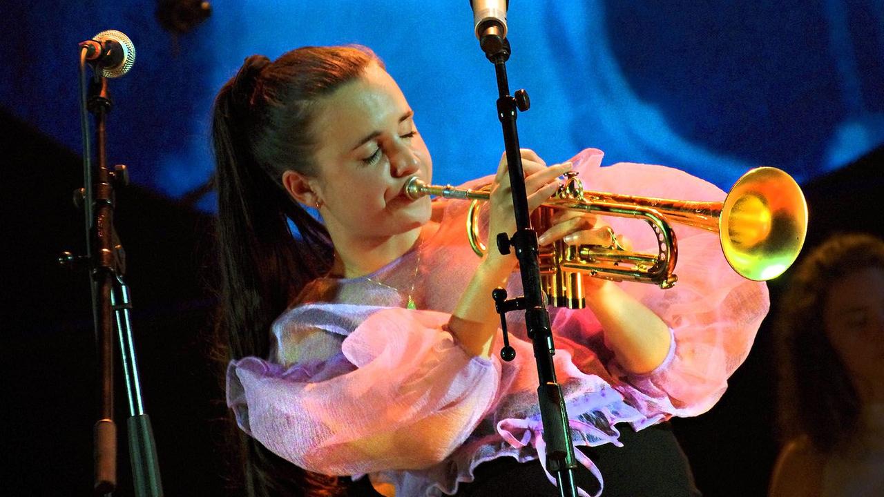 Eine junge Frau mit braunem Pferdeschwanz steht in einer Puffärmel-Bluse auf einer Bühne, von Mikrophonen umgeben, und spielt Trompete.