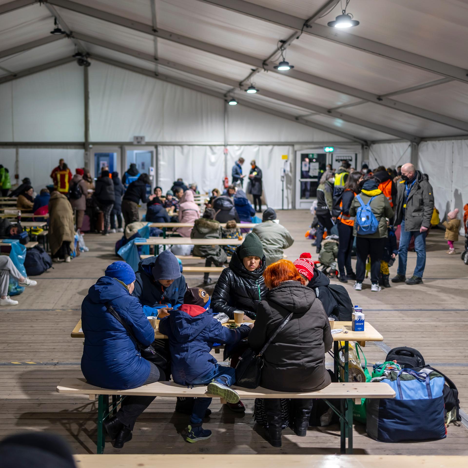 Geflüchtete in Anoraks sitzen in einem großen Zelt an Tischen auf Bierbänken.