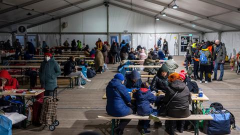 Geflüchtete in Anoraks sitzen in einem großen Zelt an Tischen auf Bierbänken.