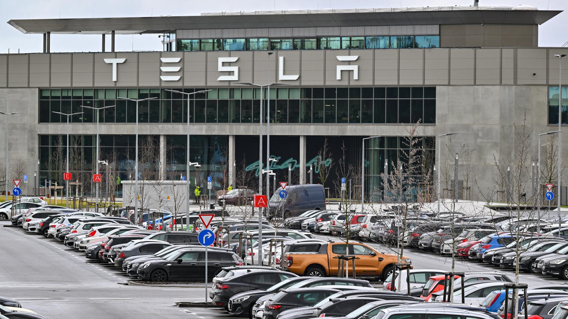 Vor einem Industriegebäude mit dem Schriftzug "Tesla" stehen zahlreiche Fahrzeuge auf einem Parkplatz.