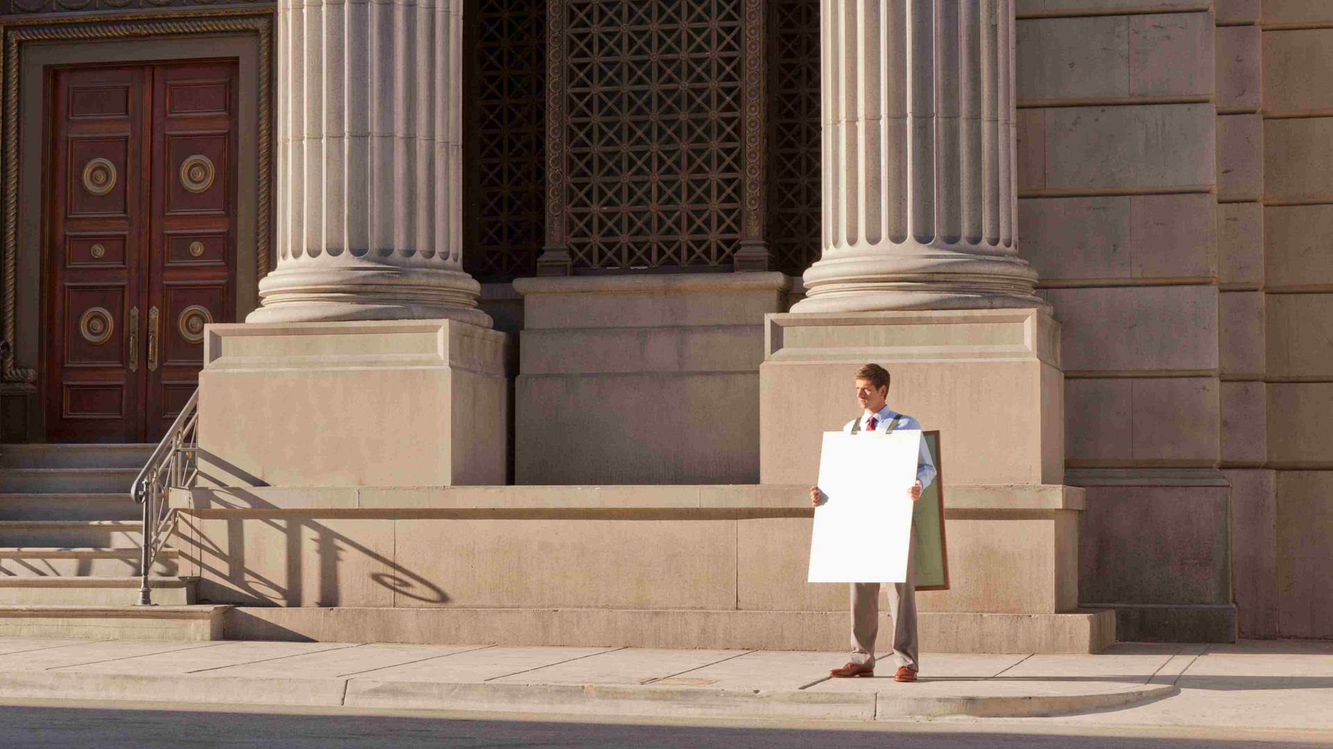 Ein Mann steht mit einem unbeschriebenen Sandwich-Board vor einem repräsentativen Gebäude mit Säulen.