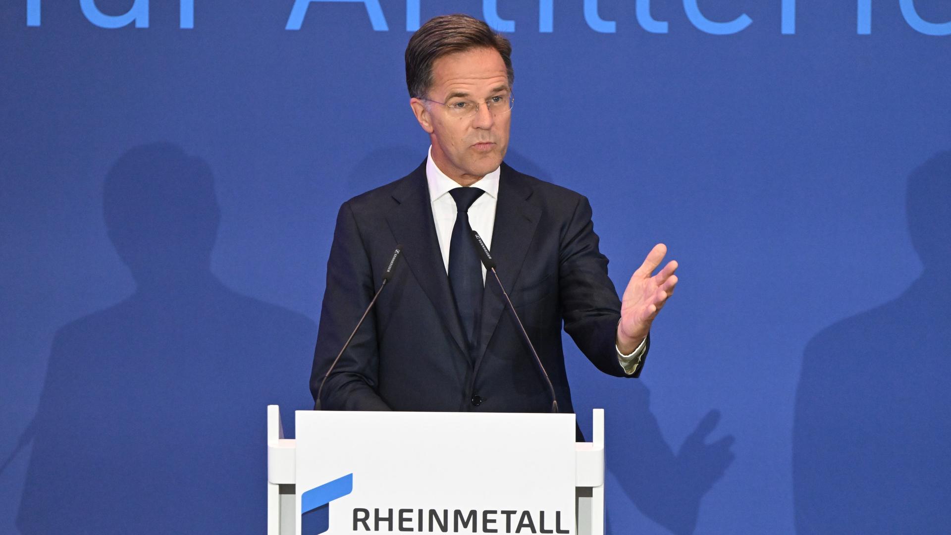 Mark Rutte steht vor einer blauen Wand an einem weißen Redepult, auf dem "Rheinmetall" zu lesen steht. Er spricht in Mikrofone und hebt seine linke Hand zu einer Geste. Mark Rutte steht vor einer blauen Wand an einem weißen Redepult, auf dem "Rheinmetall" zu lesen steht. Er spricht in Mikrofone und hebt seine linke Hand zu einer Geste.