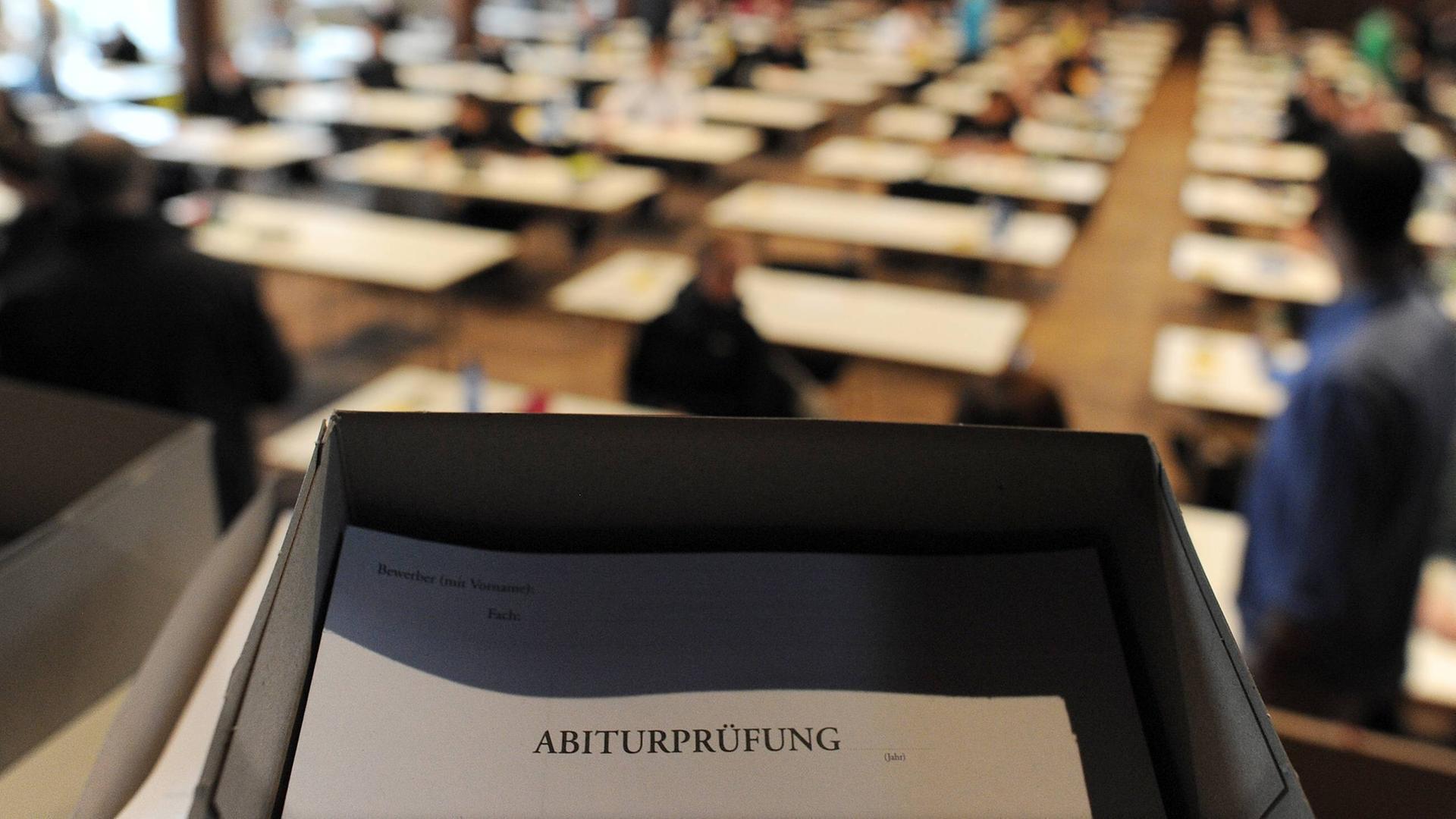 In einer Ablage liegt ein Dokument mit dem Titel Abiturprüfung. Dahinter sind Tischreihen mit Schülern zu erkennen. In einer Ablage liegt ein Dokument mit dem Titel Abiturprüfung. Dahinter sind Tischreihen mit Schülern zu erkennen.