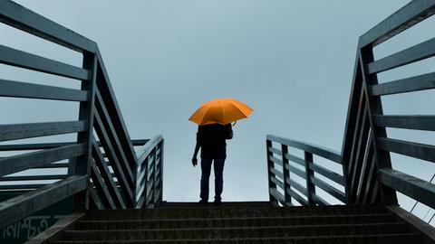 Blick auf eine große Treppe, auf der ganz oben ein Mann angelangt ist, der einen knallgelben Schirm hat, der sich leuchtend gegen den grauen Himmel abhebt.